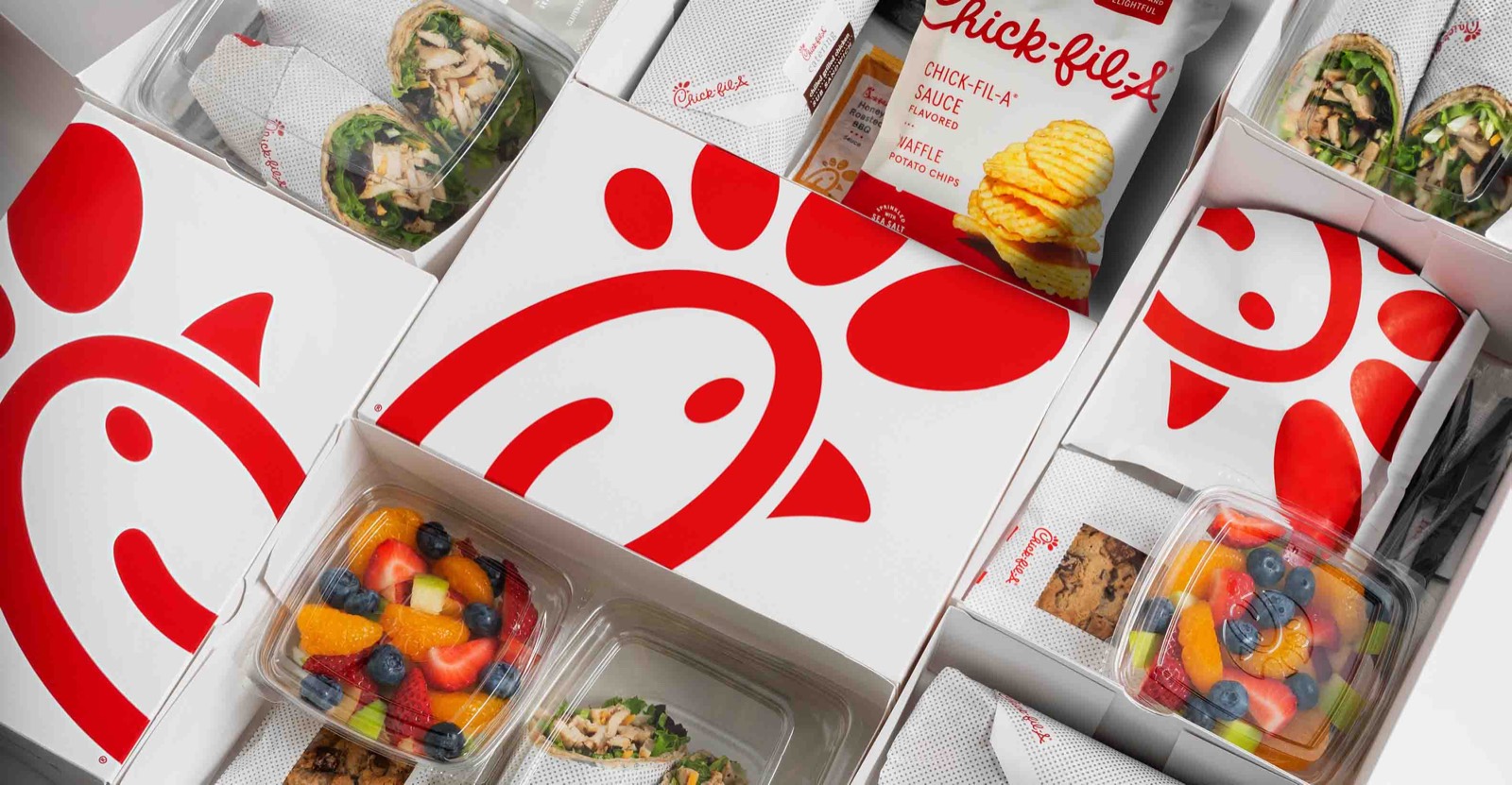 Chick-fil-A packaged catering boxes laid out with wraps, fruit cups, cookies and Waffle Potato Chips.